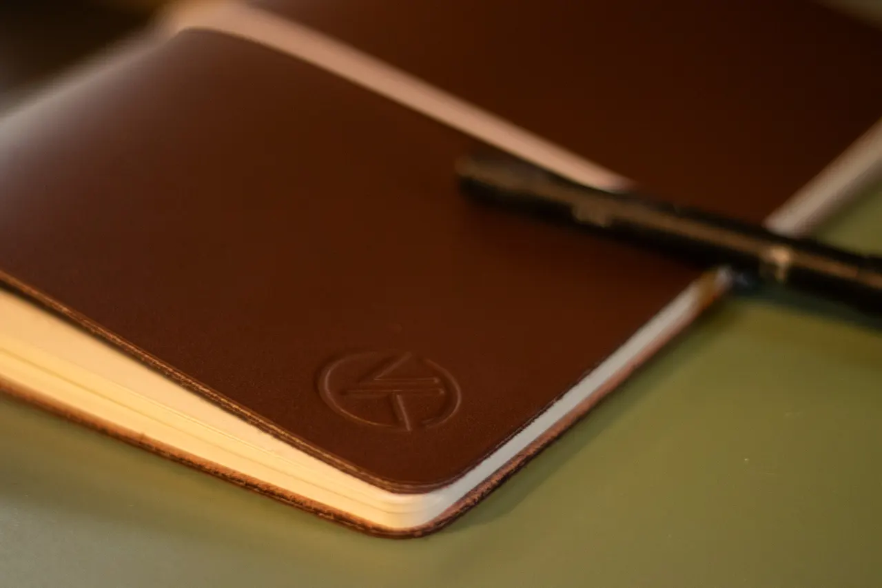 A dark brown KT Leather A5 journal lying open on a desk, the embossed KT mark on its cover and a black pen resting across the pages