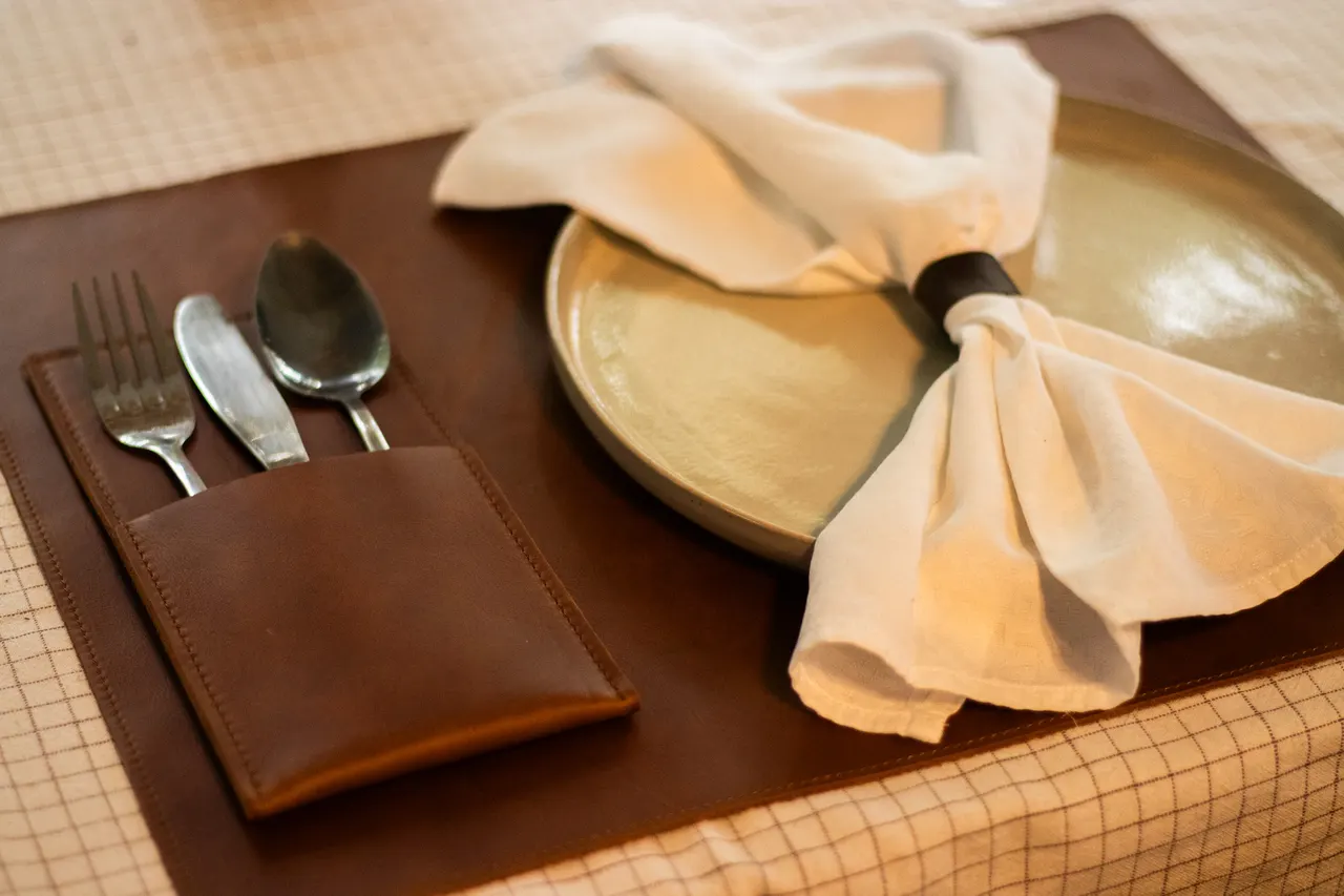 A brown leather placemat with a fork, knife and spoon tucked into its stitched cutlery pocket, set beside a plate and a linen napkin in a leather ring