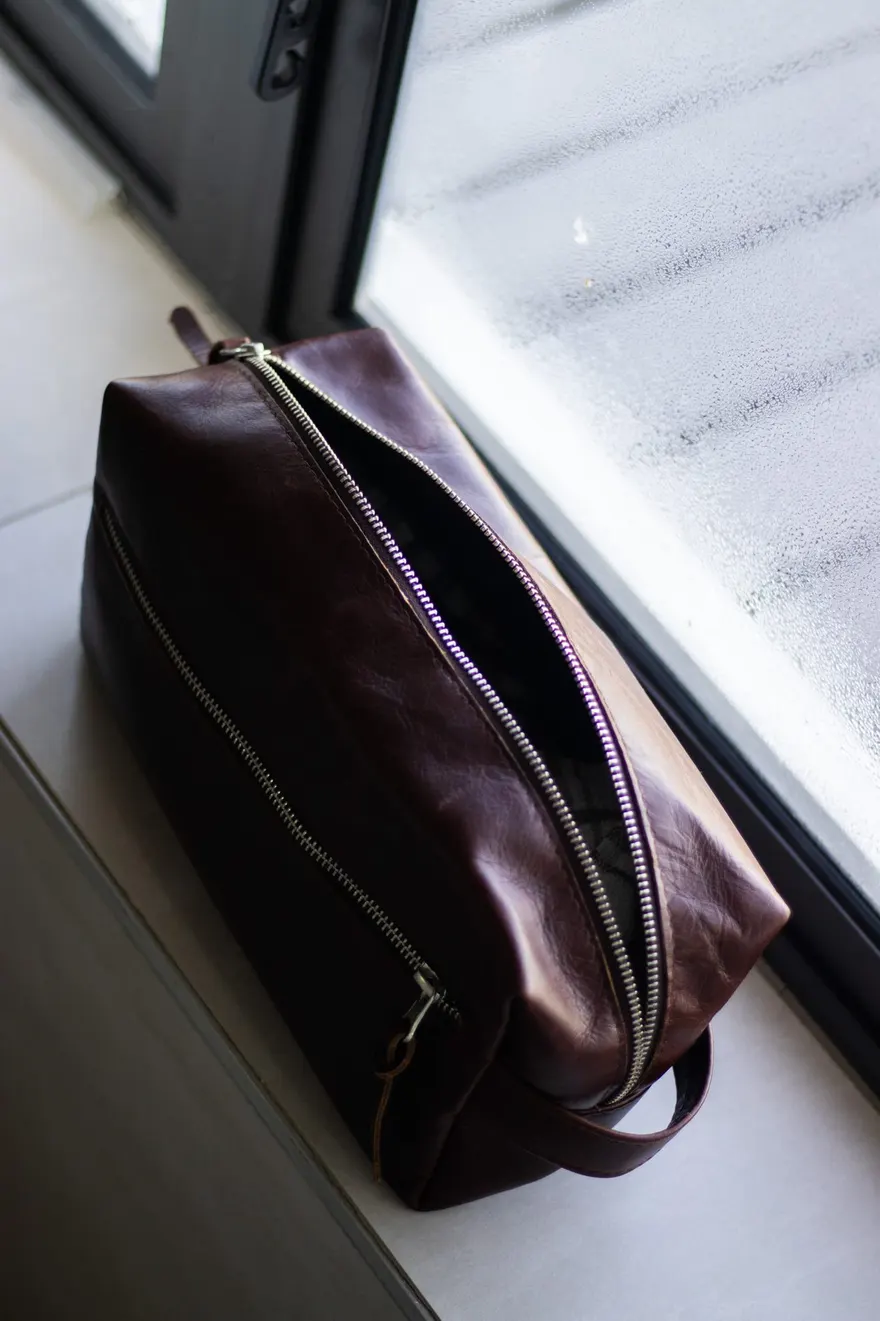 An oxblood leather toiletry bag half unzipped on a windowsill beside frosted glass